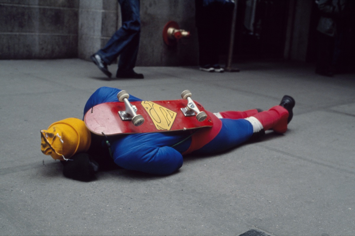 A man in a Superman suit crawling across a sidewalk with a skateboard on his back.