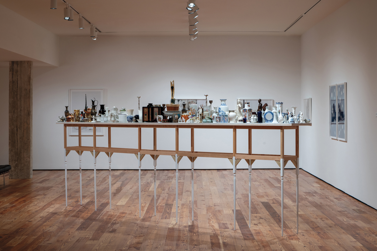 installation shot of a tall table in a gallery laden with porcelain bric-a-brac