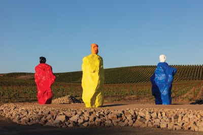A sculptural installation consisting of, from left, a large red rock with a black rock on top, a large yellow rock with an orange rock on top, and a large blue rock with a white rock on top, installed in a vineyard.