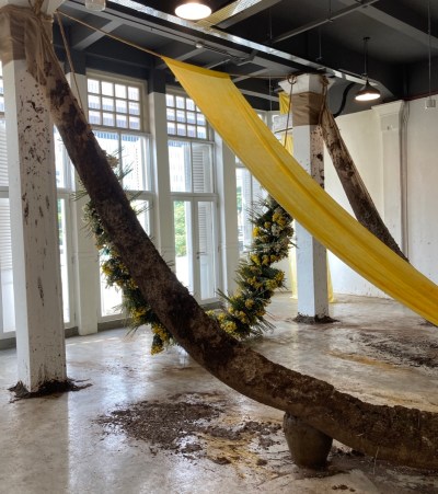 A cascading strip of yellow fabric centers an installation. In front and in back are two similar sashes but made of flowers and natural materials.