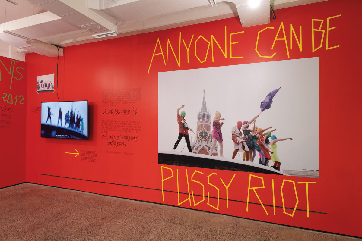 A red wall with a picture of masked activists surrounded by the words "Anyone Can Be Pussy Riot."