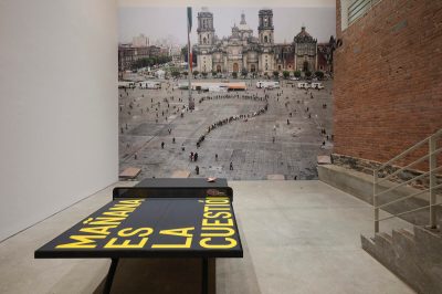 A ping-pong table painted all black, along with the phrase 'MAÑANA ES LA CUESTIÓN' in yellow. The table is set before a wall lined with a large photograph of people in a plaza arranged to form a question mark.