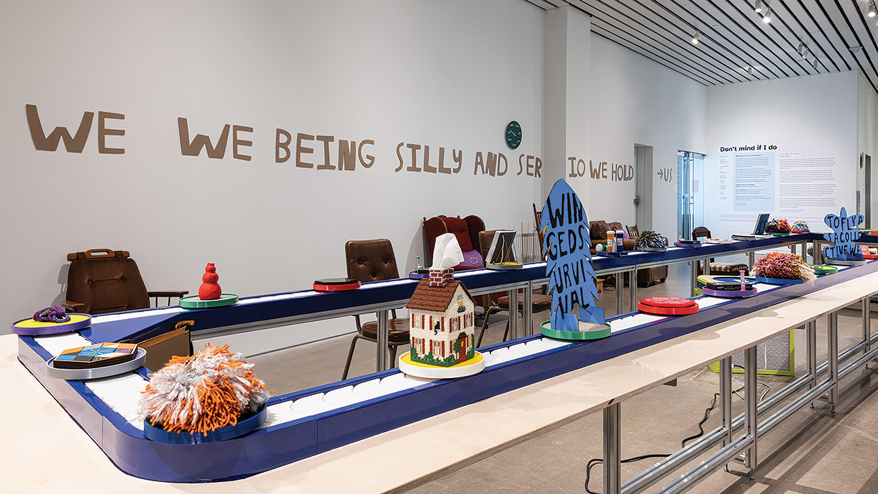 Various chairs are placed around a conveyer belt as small artworks float by. On the wall, cut-out cardboard text says "we being silly and serious... we hold..." There is a clock and wall text introducing the exhibiton, but neither are legible.