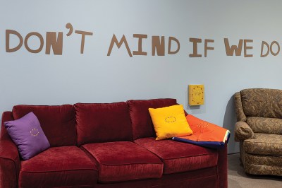 A plump red velevty couch sits under cardboard letters that spell out "Don't mind if we do." Two pillows (one yellow, one purple) and an orange blanket sit on the couch and are embroidered with a logo: arrows pointing in a loop.
