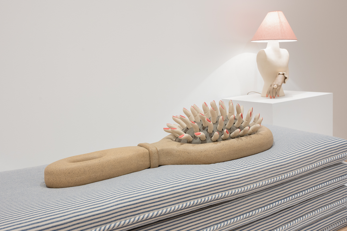 A large sculpture of a comb with human fingers for its tines. The sculpture rests on a pile of pillows. Behind the sculpture is another work resembling a turned-on lamp that sprouts a hand.