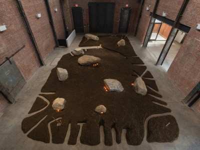 An installation of shaped dirt seen from above with rocks and lit candles set within.
