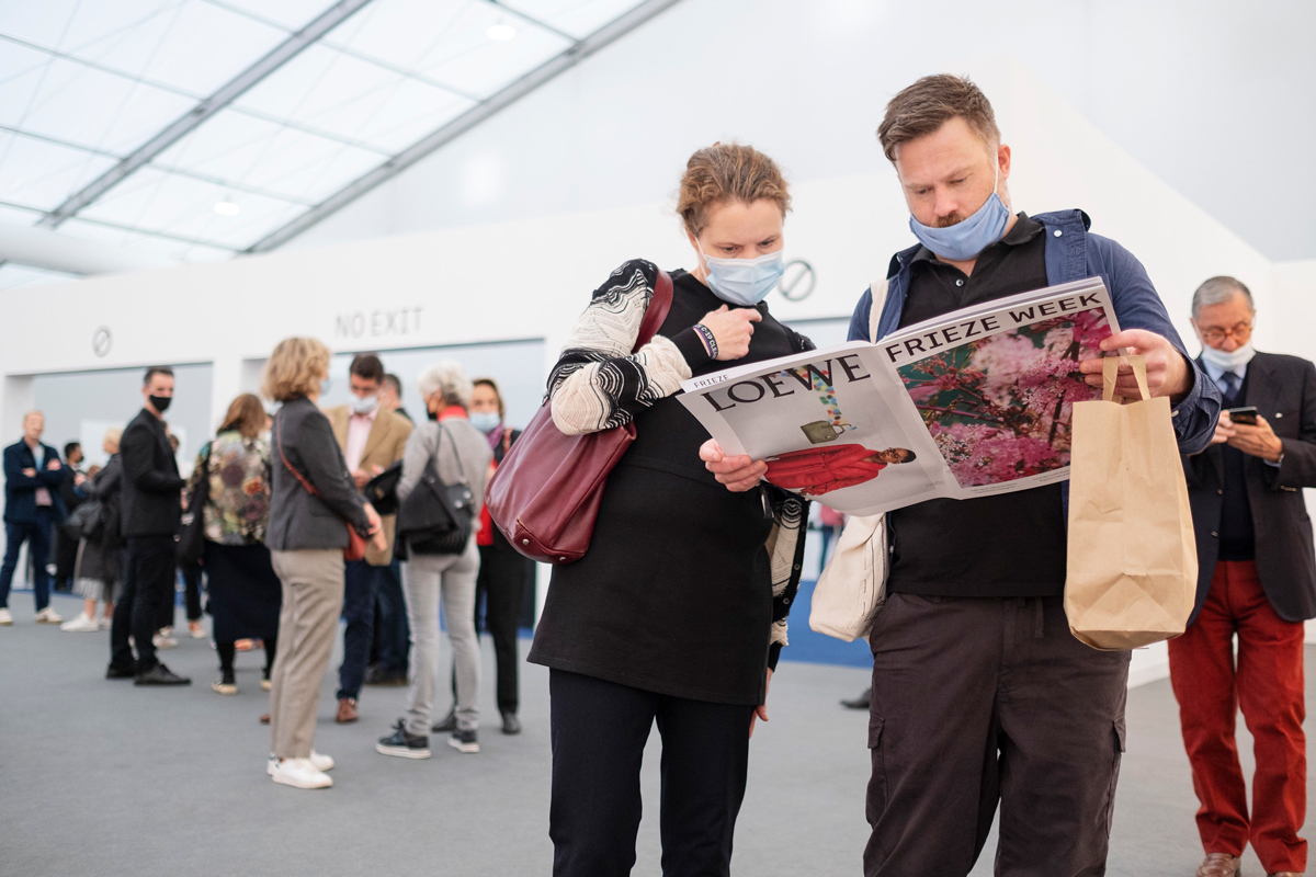 Two people look at a print publication saying 'Frieze Week' in front of a crowd of people