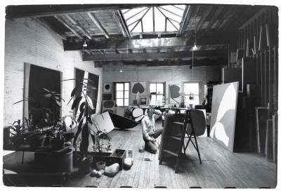 black and white photo of a loft-like space full of art and plants, with a man kneeling down in front of a canvas