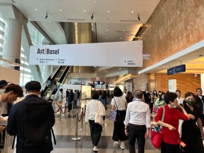 A crowd of people walkthrough a convention center lobby with a sign overhead that reads 'Art Basel'.