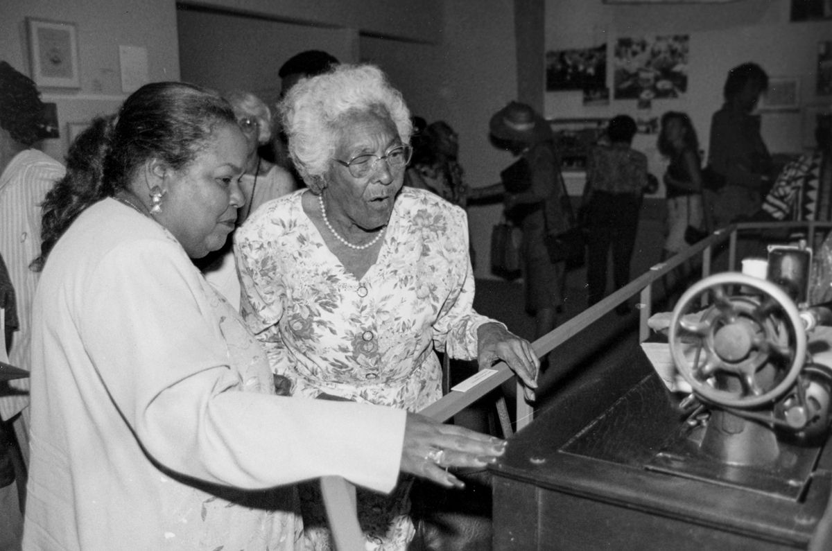 Archival photograph of two Black women looking at a sewing machine on view in a museum.