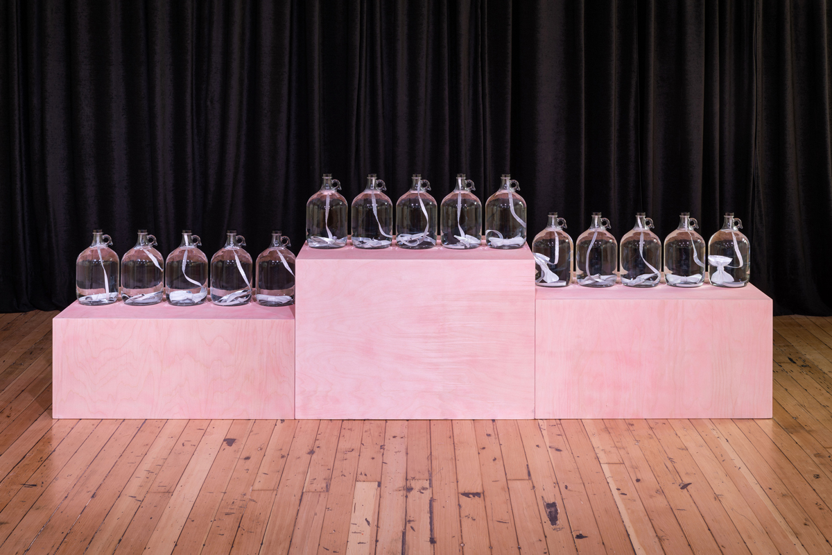 Three award-ceremony-like platforms in pink hold five glass jugs filled with a white ribbon.