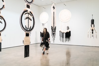 A man wearing a face covering, a black robe, black bands, and a black sock walks through a gallery with hanging fiber sculptures.
