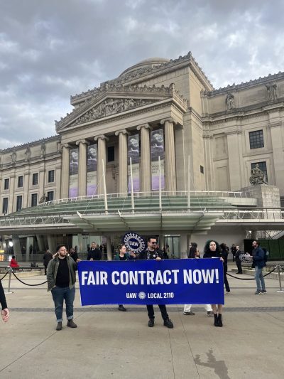 People hold a blue sign that reads FAIR CONTRACT NOW! in front of the Brooklyn Museum.