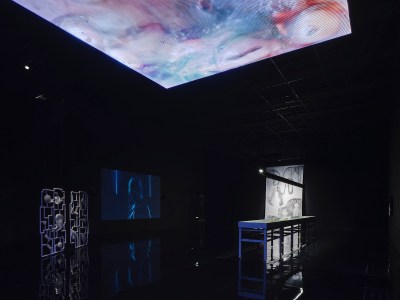 A ceiling hung with a screen displaying an abstract pattern that illuminates a mostly darkened room. A sculpture resembling a gate and a video playing on a screen are barely visible within.
