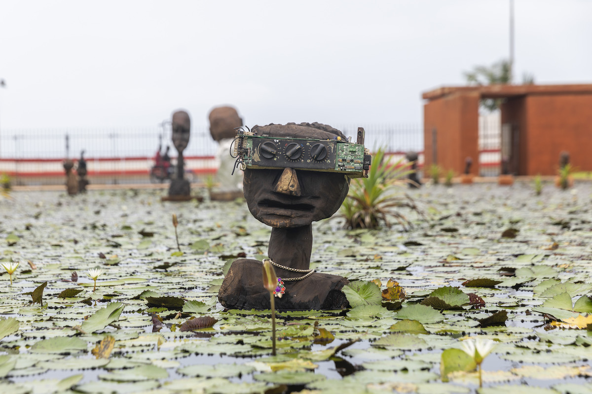 A sculpture of a Black man's head with a radio covering his eyes. The sculpture is set within a pond covered in lily pads.