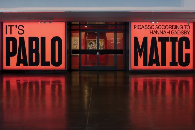 A giant read sign reading IT'S PABLO-MATIC: PICASSO ACCORDING TO HANNAH GADSBY at the entrance to an art gallery. A painting is visible behind its doors.