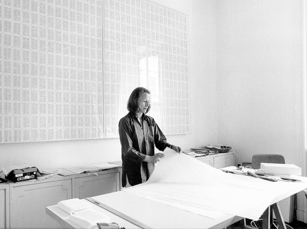 A black-and-white image of a white woman picking up a large sheet of paper that's placed on a drawing board. Behind her is a two-panel work that resembles a series of scribbles arranged in grids.