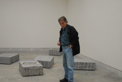 A white man in jeans, a button down shirt, and a black jacket standing amid a room filled with rough-hewn stones on its floor.