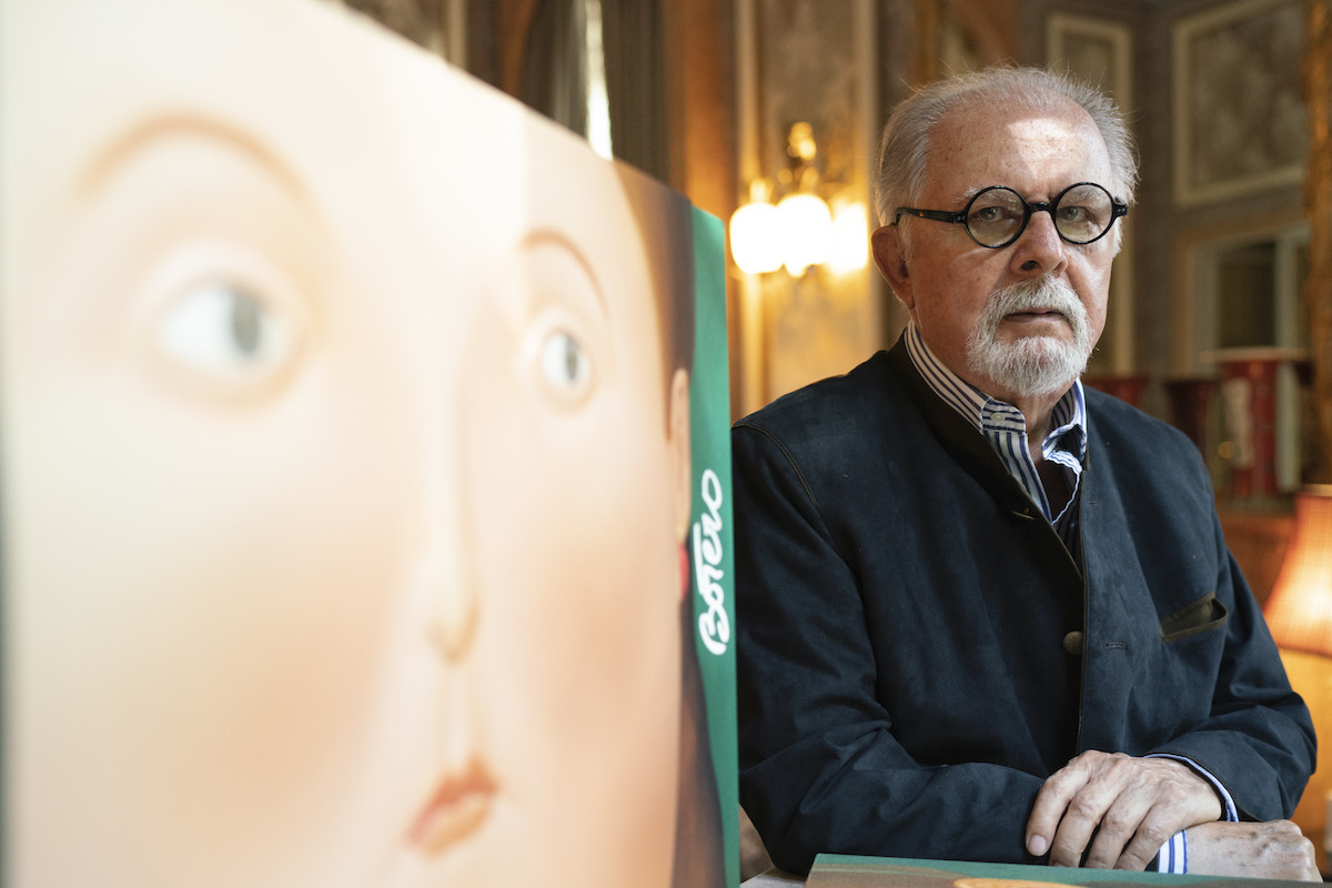 A man in glasses with a beard standing next to a painting of a large woman's face.