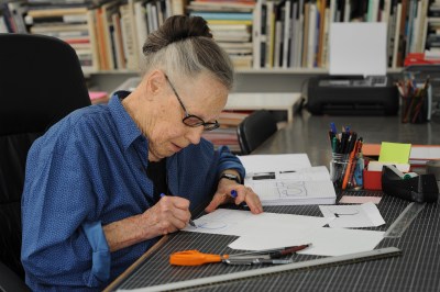A woman with her graying hair in a bun drawing on a sheet of paper. A cup with pens and a pair of scissors sit beside her. Behind her is a shelf filled with books.