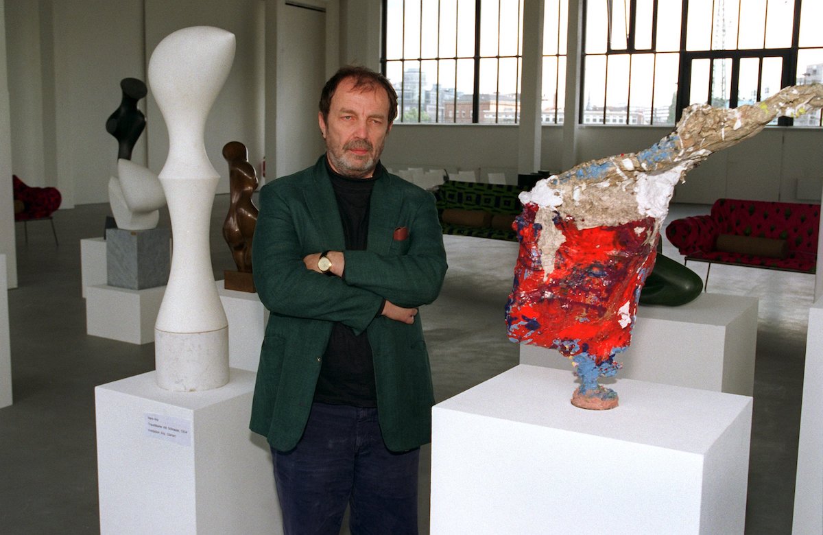A white man in a green suit jacket standing with his arms crossed beside two abstract sculptures on plinths.