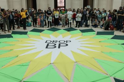 A group of people in a large hall stand around a bright green and yellow banner that says DROP BP in the style of the BP logo, which resembled an angular sun or flower.