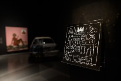 A black album cover showing a crown, phrases such as 'TEST PRESSING,' and more scrawled in a cartoonish style. It is set in a gallery in which, in the background, out of focus, is visible a car and a painting of a Black woman.