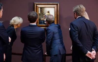A group of people in suit jackets staring at a painting of a woman pouring milk from a vessel.