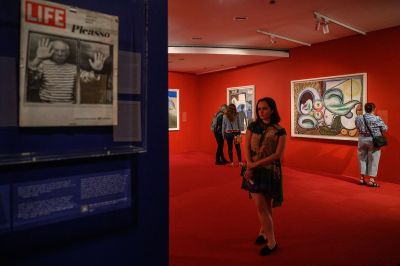 A woman standing with her legs crossed in a red gallery whose walls are lined with paintings. Nearby is a LIFE magazine cover with a man in a Breton shirt holding his hands up to a pane of glass.