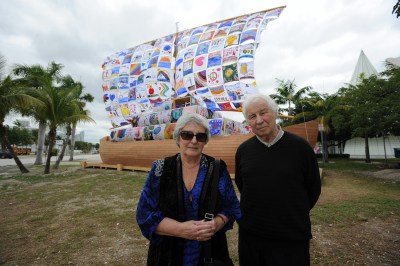 A white woman and a white man standing before a ship whose sails are lined with children's drawings. The ship is placed on land amid palm trees.
