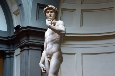A color photograph shows a large, tall, white sculpture: a handsome man. He is nude, and he stands with his left arm partially raised in an ornate domed space.