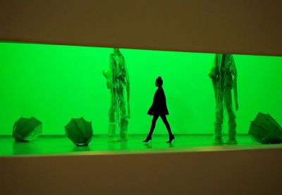 A woman walking through a gallery lit a bright shade of green. She walks beneath two sculptures of towering figures draped in fabric. Around them are objects resembling inverted umbrellas.