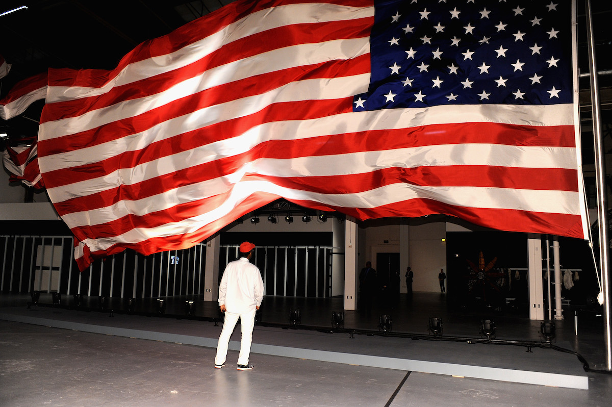 A Black man in a red cap standing beneath a gigantic American flag blowing in the wind.