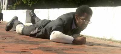 A Black man in a suit crawling on his hands and knees across a brick pathway. He has padding on his arms and kneecaps.