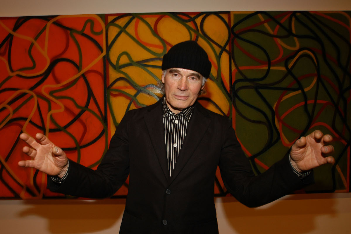 A man in a black beanie and suit jacket with his arms outstretched. He stands before a large abstraction.