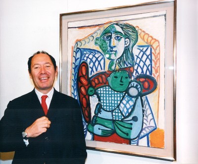 A smiling man in a suit beside an abstracted painting of a woman with a baby.