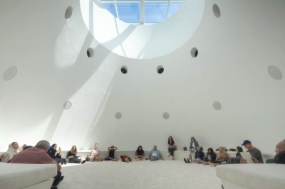 A number of people sit on white carpet in a domed, white space featuring speakers embedded into the wall.