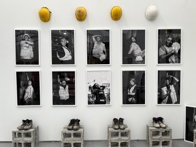 An installation featuring two rows of photographs in the center, a row of construction hard hats above, and boots on cinder blocks below.
