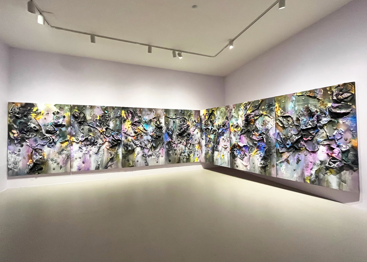 An abstract painting with various things collaged onto the surfaces takes over two walls in an art museum. 