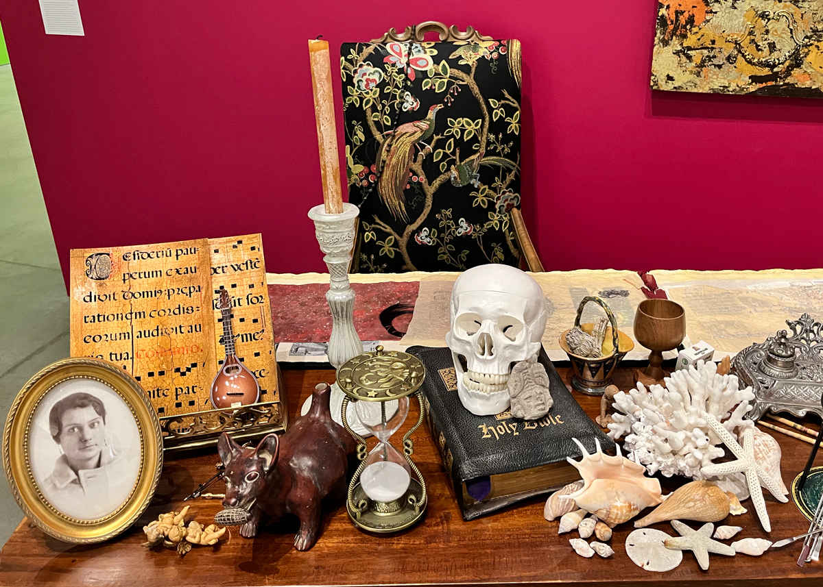View of an art installation composed of various objects, including a candle, skull, hourglass, bible, shells, and photograph.