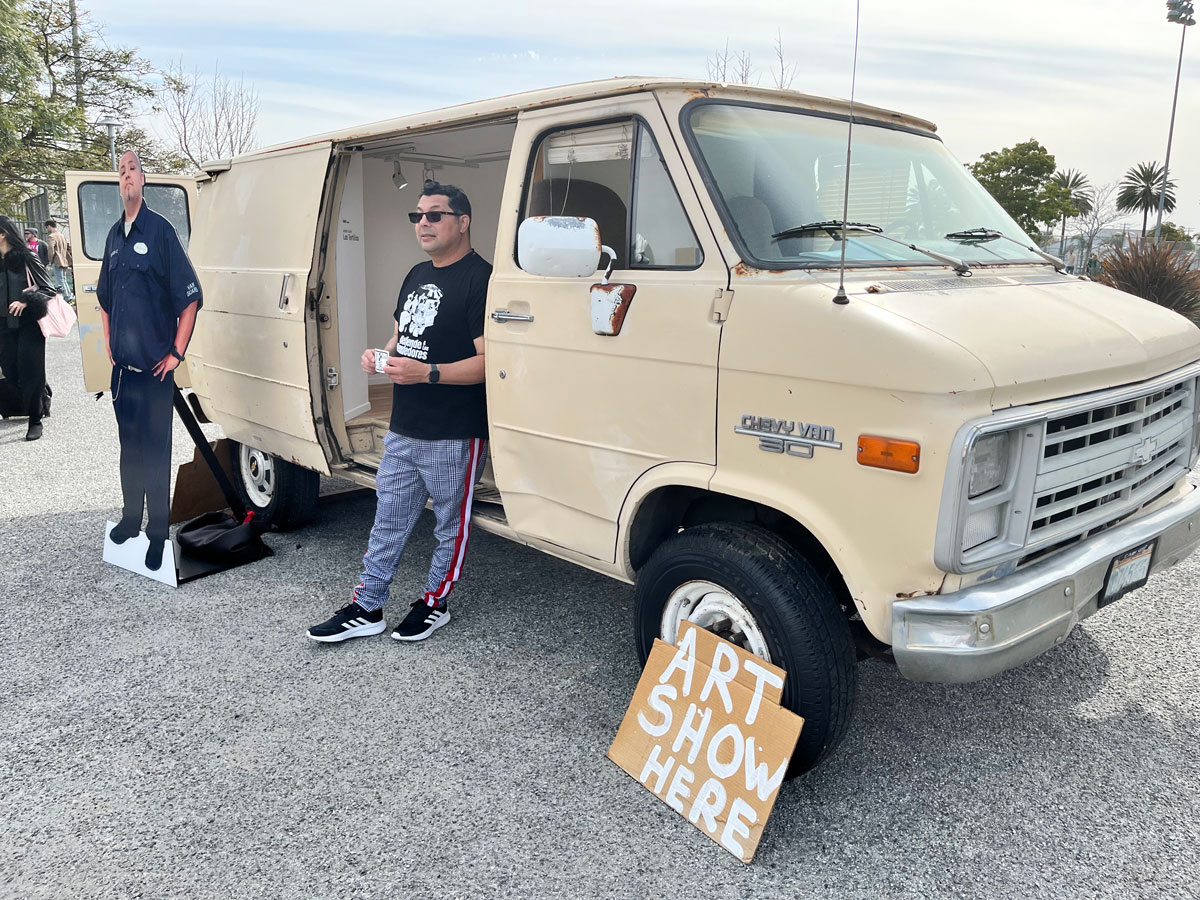 Ruben Ochoa stands in front of a beige van that is also an art gallery. 