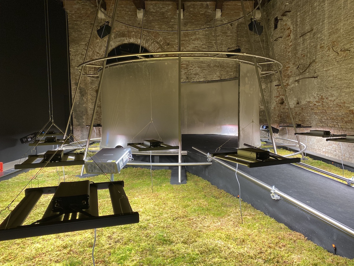 A round screen at the top of a ramp surrounded by large projectors set in moss.