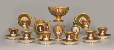 A group of teacups and bowls, as well as a kettle, with painted images of white women's faces on them. The teacups and bowls are ornate, with the cups giving gold and green plates that match them.