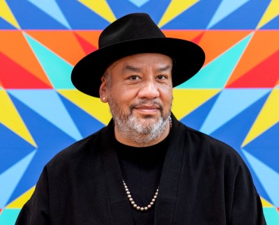 A Brown man wearing a black hat and jacket, along with a necklace. He stands before a vibrantly colored abstraction.