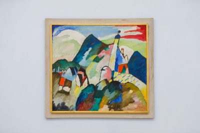 A square painting with vibrant (and unusual) colors, showing a mountainous landscape, hangs on a white wall in a photograph.