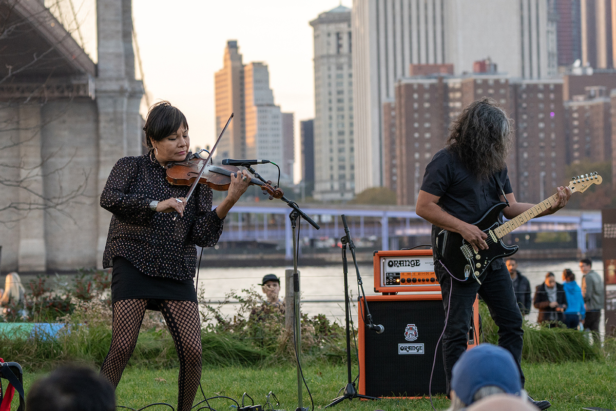 A woman in black clothes playing a violin by the side of a black-clad guitarist, both in front of the Brooklyn Bridge.