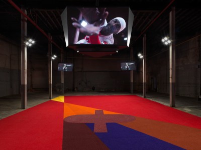 A long carpet with an oval bisected by a rectangle with bright lights shining down on it. Above, a large screen shows a Black man in football gear bringing his hands to the camera.