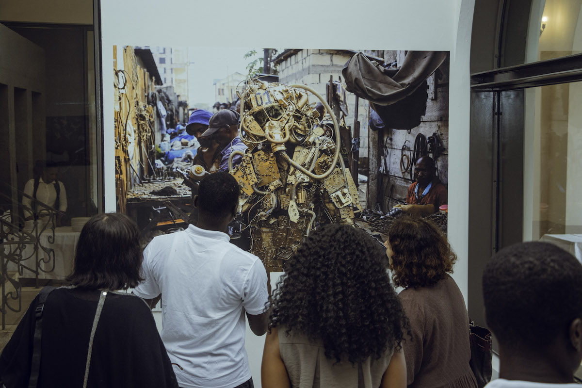 A group of people staring at a photograph of a person in a market wearing machine parts that have been painted gold.