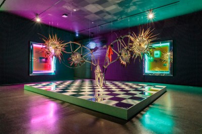 A hanging sculpture whose ends have sunburst-like forms that sits atop a raised checkerboard floor. On the walls nearby are paintings with neon pink and green lighting.