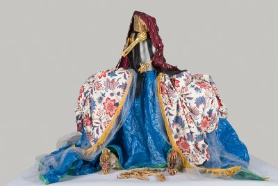 A sculpture resembling a person whose dress is split open to reveal tarp and animals' heads. The faceless person holds one gold arm up.